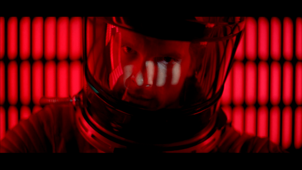 Screencapture from 2001: A Space Odyssey. In a red room, a man in a space suit is looking directly at the camera.