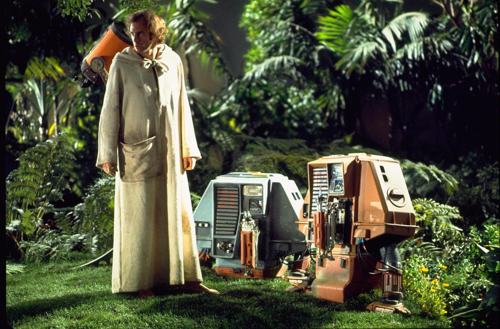 Screencapture from Silent Running. A man stands in a forest wearing a robe next to two small robots.