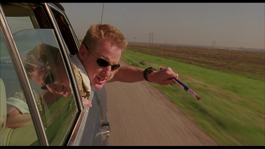 Screencapture from Bottle Rocket. A man fires a firework from the window of a moving car. 
