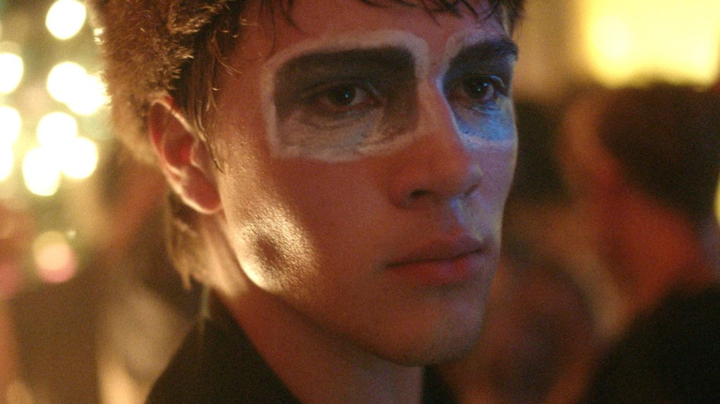 Oscar is wearing blue and white paint around his eyes with bright lights behind him.