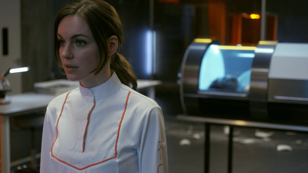 A woman, in a white futuristic suit, stands in a laboratory. From the film The Complex.