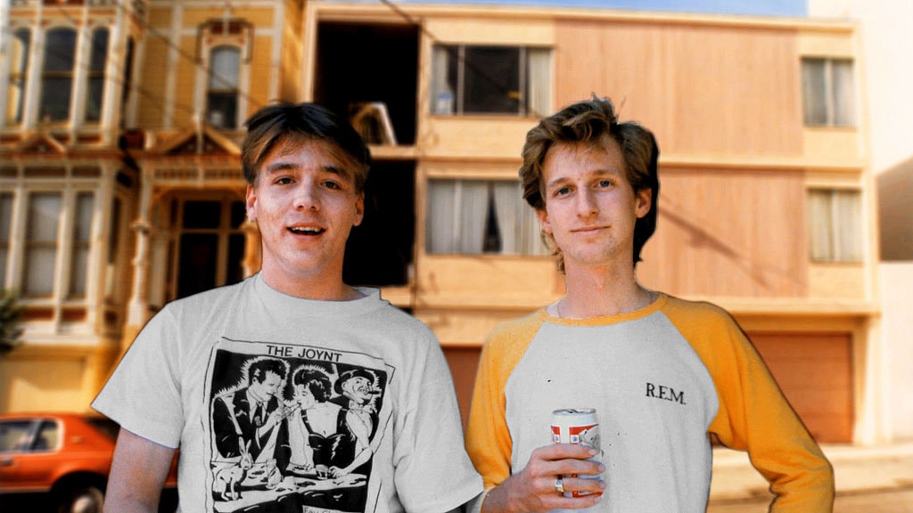 Two young men in band t-shirts stand in front of a run-down pink apartment building. The two men are Mitchell D. and Eddie Lee Sausage, respectively.