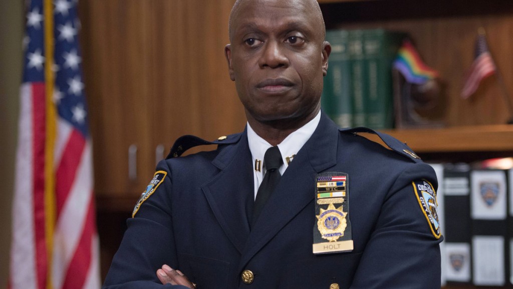 Captain Holt wears his police uniform and stands in his office with his arms folded.