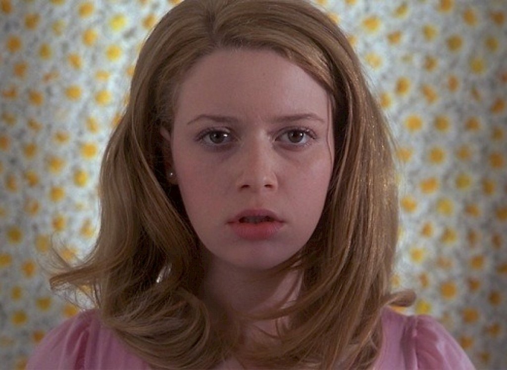 Dressed in a pink, Megan stares straight at the camera as flowers cover the wall behind her.