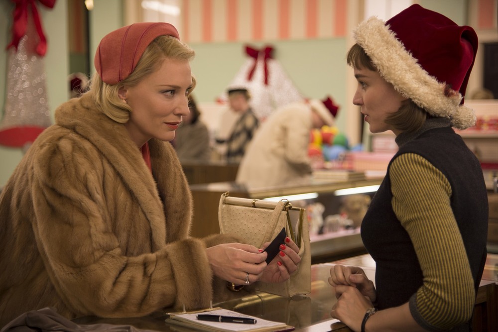 Carol, who is wearing a fur coat, leans over the store counter to talk to Therese, who is wearing a Santa Hat.