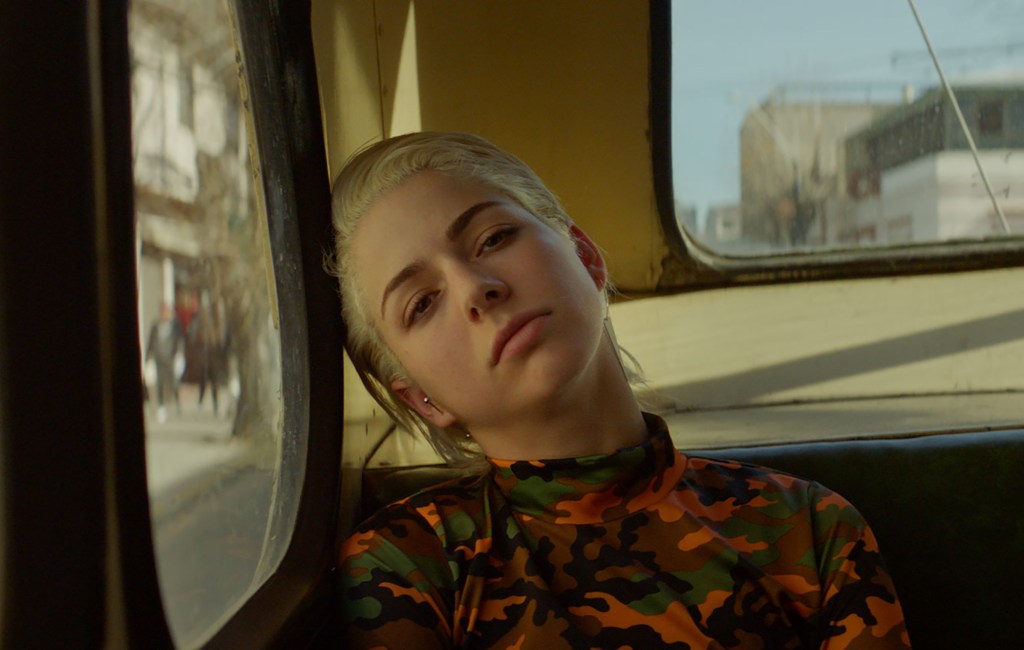 Ema rests her head on the window of a bus. She is wearing a read camouflage turtleneck.