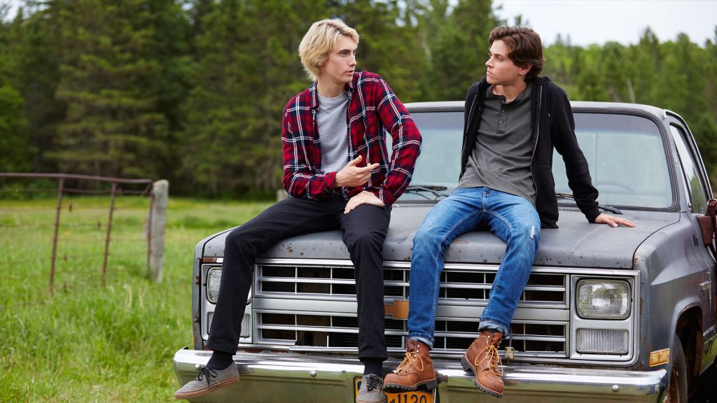 Lukas and Philip sit on the hood of a grey truck in the countryside. They are engaged in coversation.