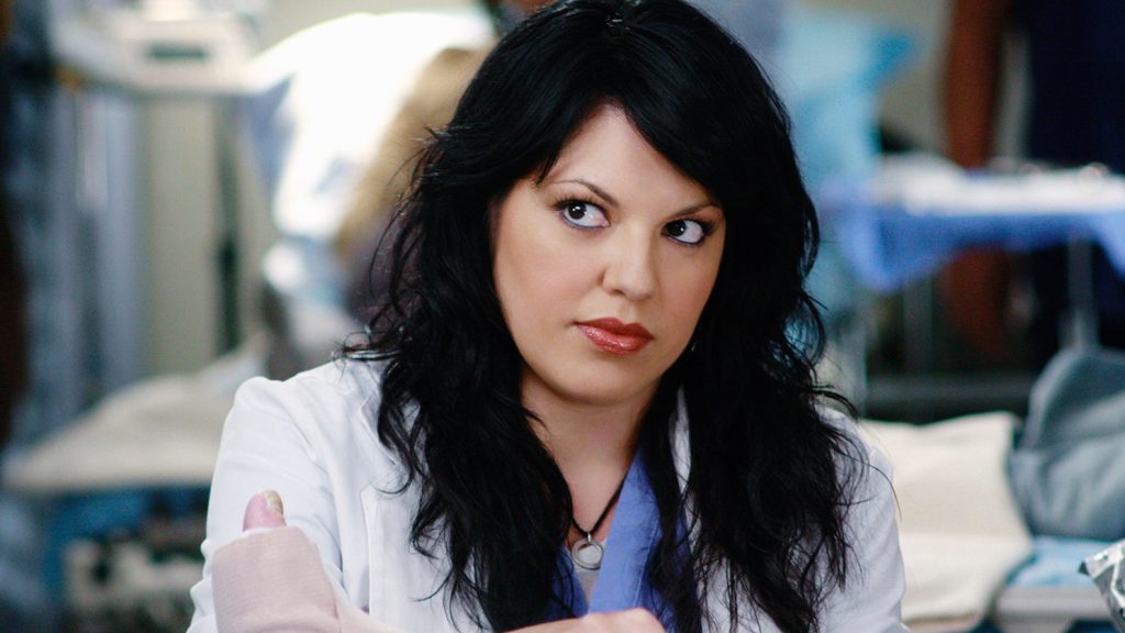 Callie is dressed in a lab coat and scrubs as she works on a patient.
