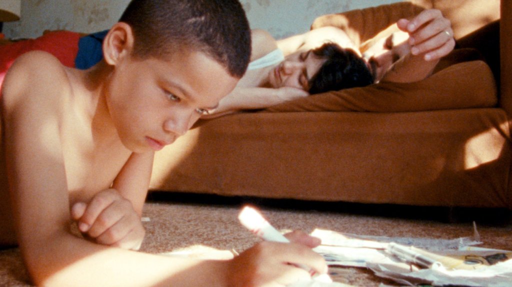 Jonah lays on the floor and colours. Two adults are asleep on the bed behind him.