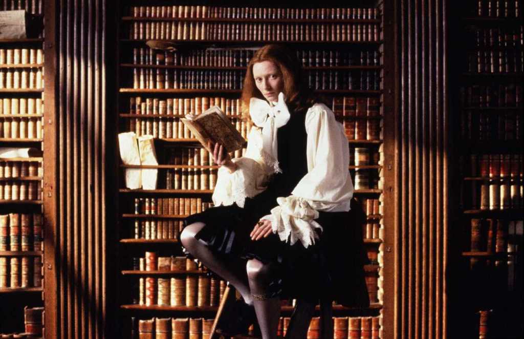 Orlando is reading a book in a library. They are dressed in flamboyant period clothing, with a large white bow and puffed sleeves.