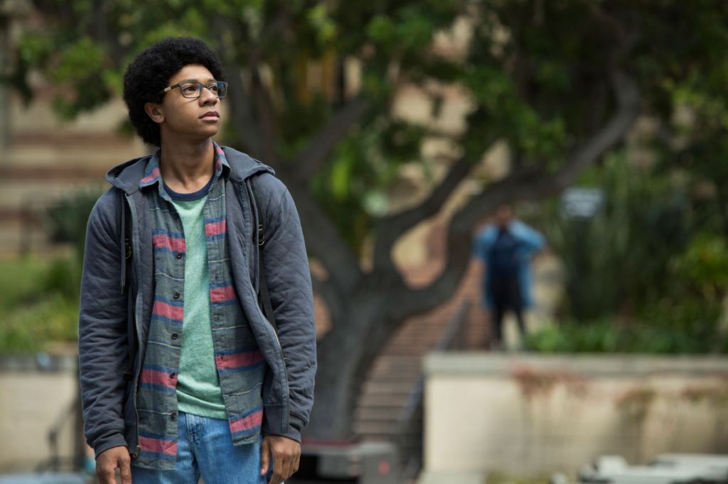 Lionel walks around his college campus. He dressed in layers of dark clothing with a pale green shirt at the bottom.