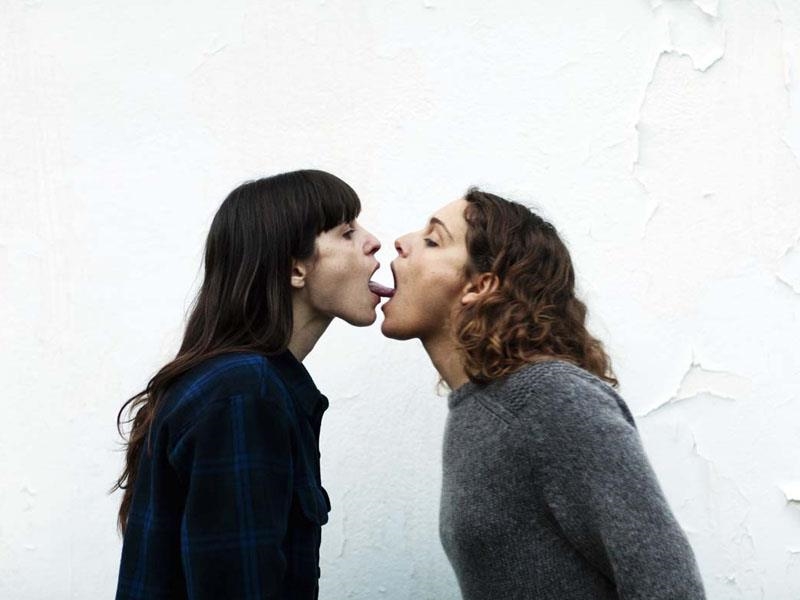 Two women stand facing each other, leaning in as their tongues reach into each others' mouths. 