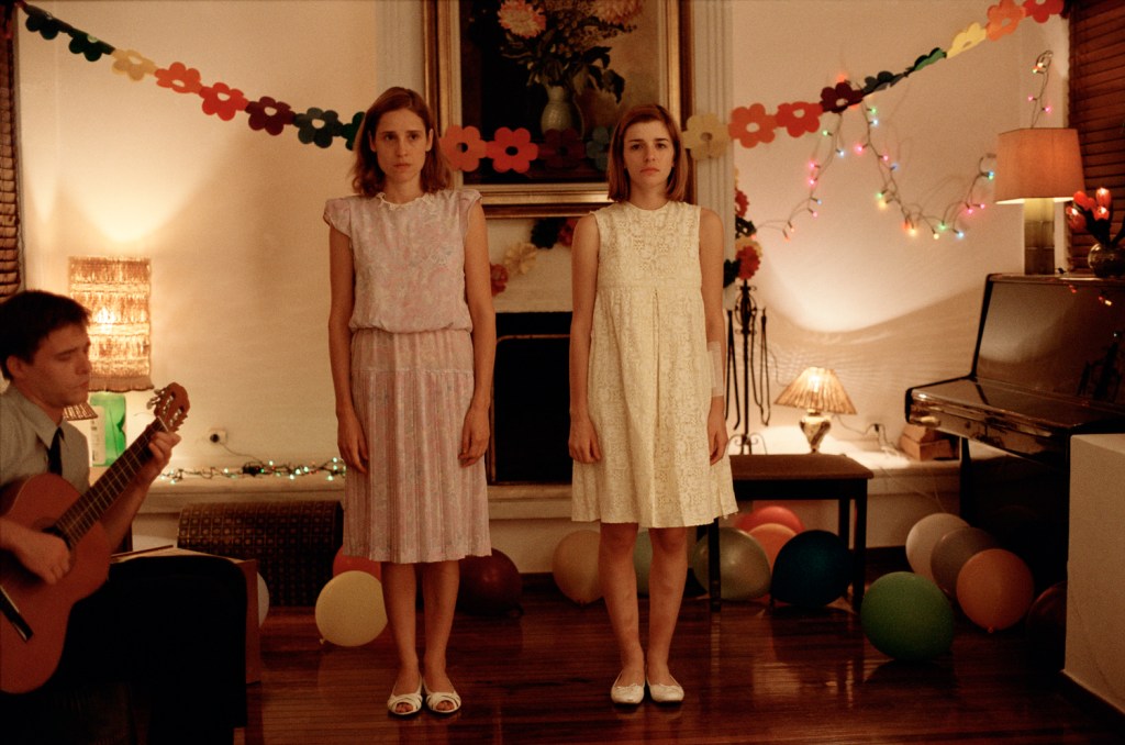 Still from 'Dogtooth'. Features two girls stood quite rigidly, looking unhappy. A man on the left sits and plays guitar. There are party decorations in the room.