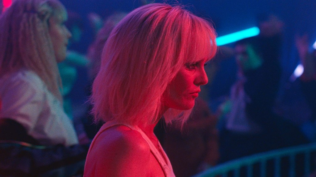 This image is from the film Knife + Heart. A woman stands in the middle of the image, with a red light covering her. She is in a nightclub, which is entirely blue.