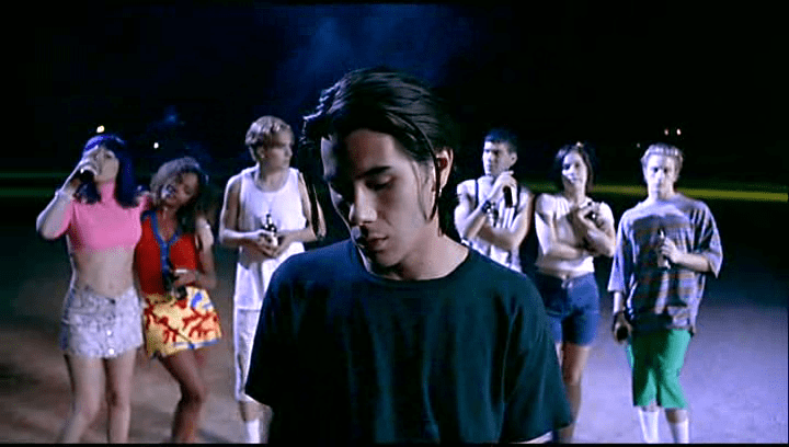 Image is from the film Nowhere (1997). A man looks down in the center of the shot. A series of teenagers stand behind him drinking beer.