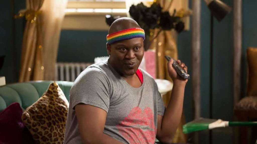 Titus pouts whilst holding a TV remote in his hand. He is wearing a rainbow coloured headband.