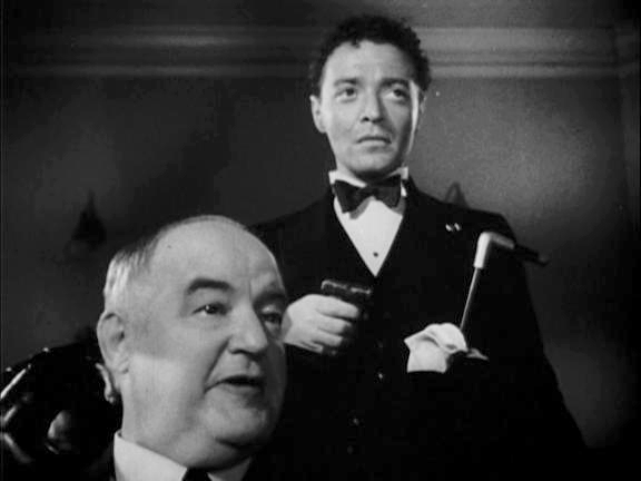Joel Cairo [Peter Lorre] is holding a cane and is standing behind Kasper Gutman [Sydney Greenstreet]. Both are looking off screen and wear stern and displeased expressions