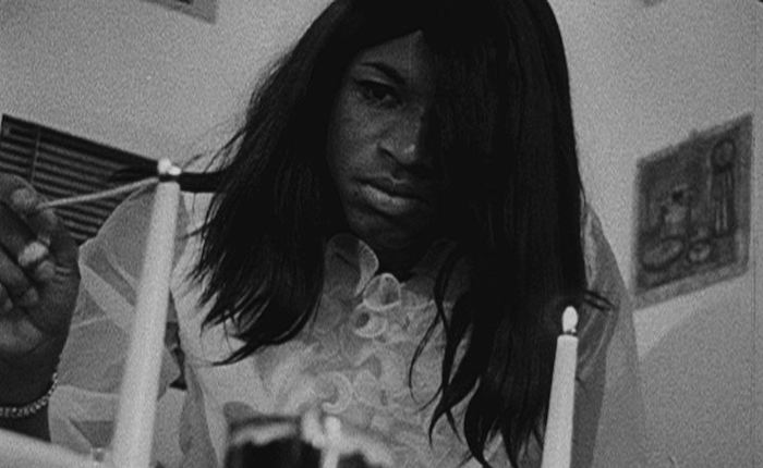 An unnamed Black woman stands above the camera, wearing a neutral expression. She wears a white frilly blouse and is lighting a pair of thin white candles