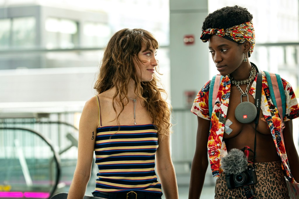 Ash and Honeybear walk side by side, looking at one another. Ash is wearing a striped tank top whilst Honybear wear an open shirt with nothing but nipple tape underneath.