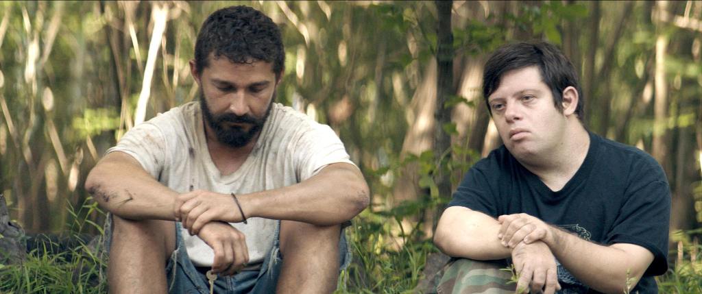 This is a still from the film Peanut Butter Falcons. Actor Shia LaBeouf is sitting down on the ground with actor Zack Gottsagen. There is a background of tree.
