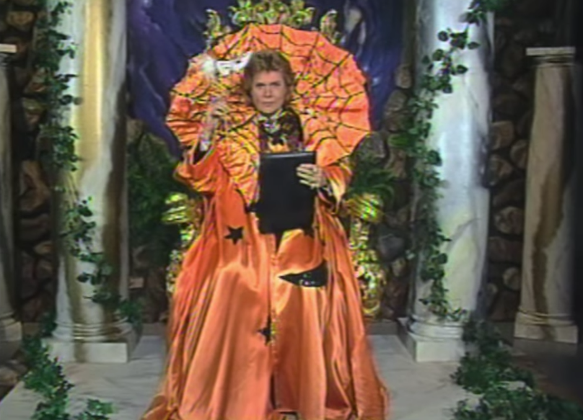 An old image from Walter Mercado's television program. Walter reads from a large book dressed in a huge orange cape decorated for Halloween. He looks like a large pumpkin.