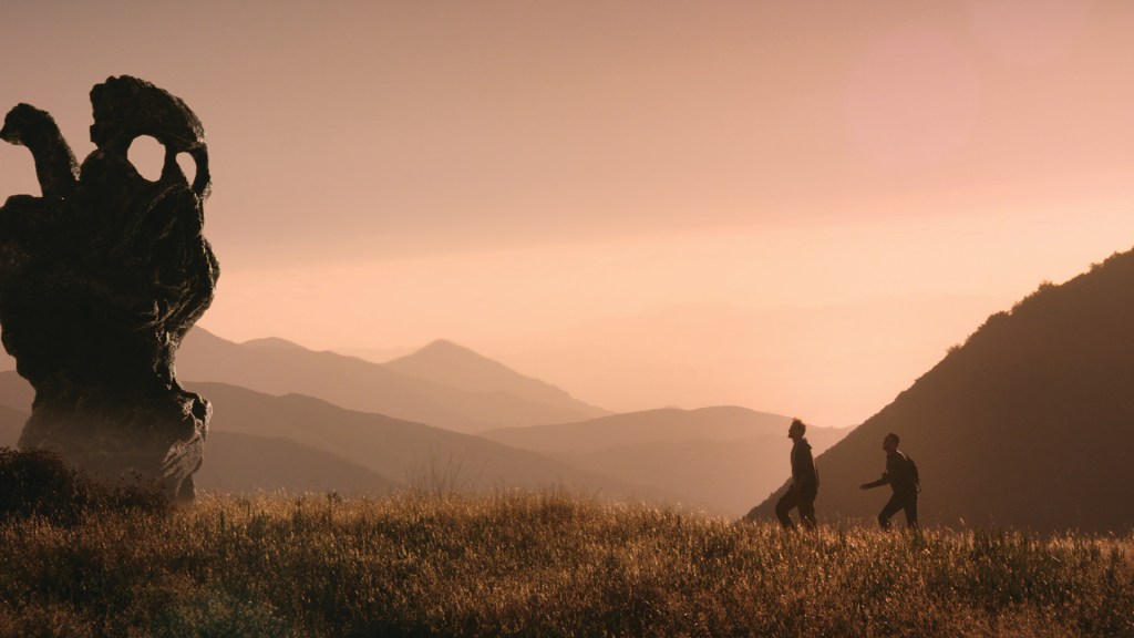 This image is from The Endless. Two men walk through a meadow and approach a skull-like statue. 