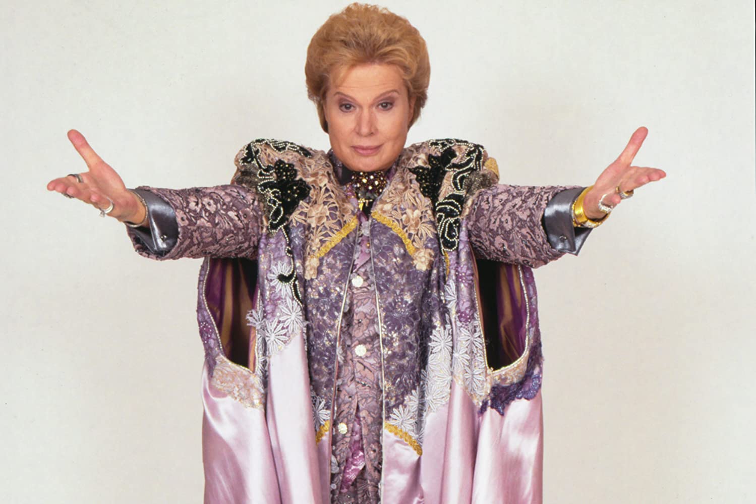 Walter Mercado stands arms spread out presenting himself. He wears an extravagant purple robe with intricate design.
