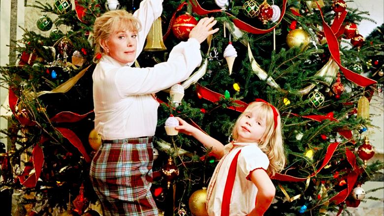 Nanny and Eloise stand in front of a massive Christmas tree decorated with ribbon, sphere ornaments, and cupcake ornaments