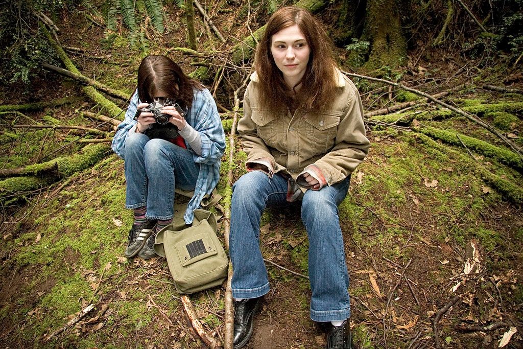 Image from We Go Way Back. Young Kate and Older Kate sit on a small hill in a forest. Young Kate is taking pictures, whilst Older Kate is watching something in the distance.