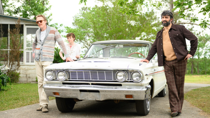 Three people stand next to an old car. Two have their hands on the bonnet.