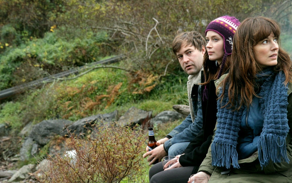 Image from Your Sister's Sister. Jack, Iris and Hannah sit in the countryside with a few drinks. Jack is looking at Hannah, who is looking behind the group at something unseen. Iris, on the other hand, looks ahead as if unaware of the other two.