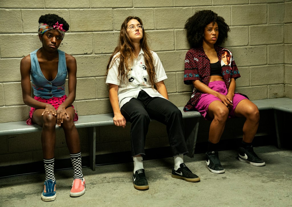 Janay, Honeybear and Camille sit with their backs against a white brick wall. They are in a jail cell. 