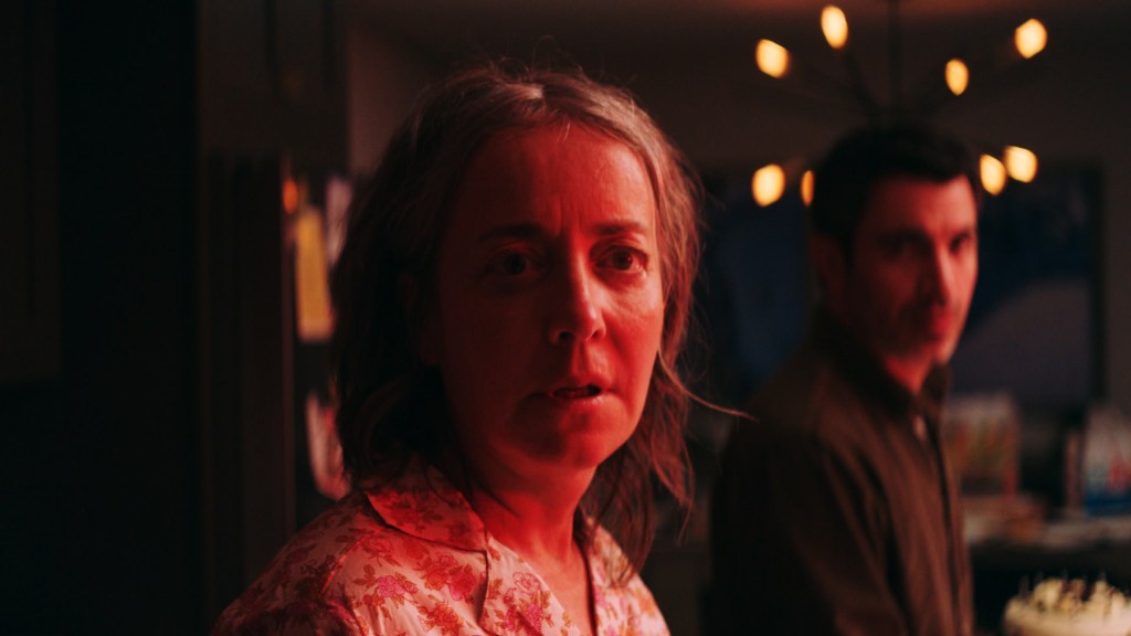 Jane (Jane Adams) in her pajamas, bathed in red light. Jason (Chris Messina) stands behind her.