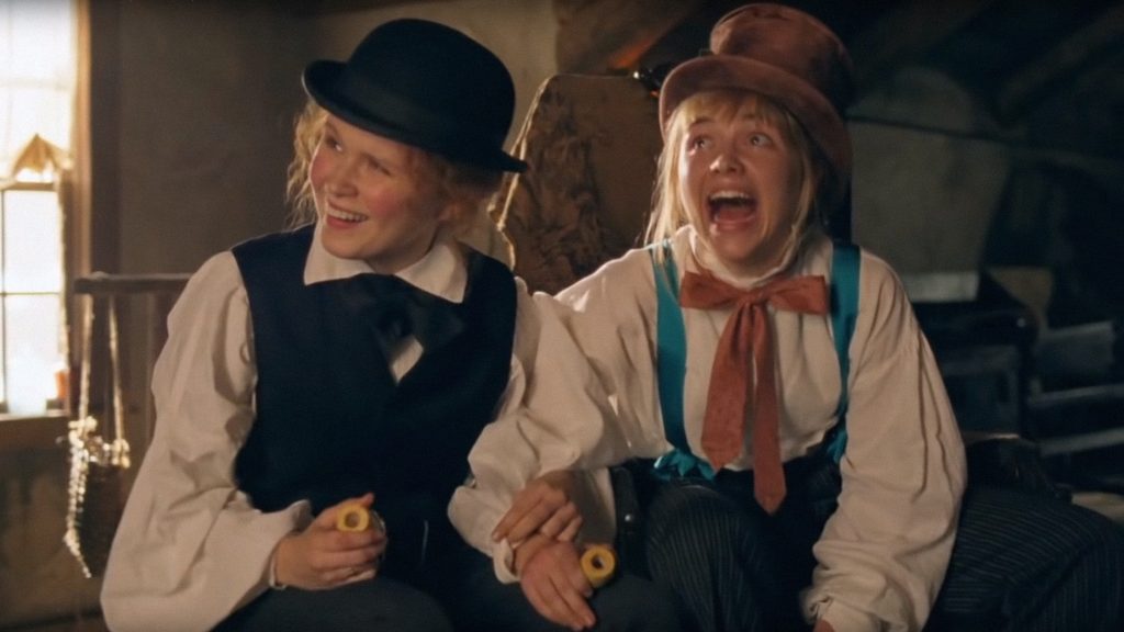 Image from Little Women. Two girls play together whilst dressed in men's clothes. One is wearing darker clothes whilst the other wears blue and brown, and appears to be shouting.