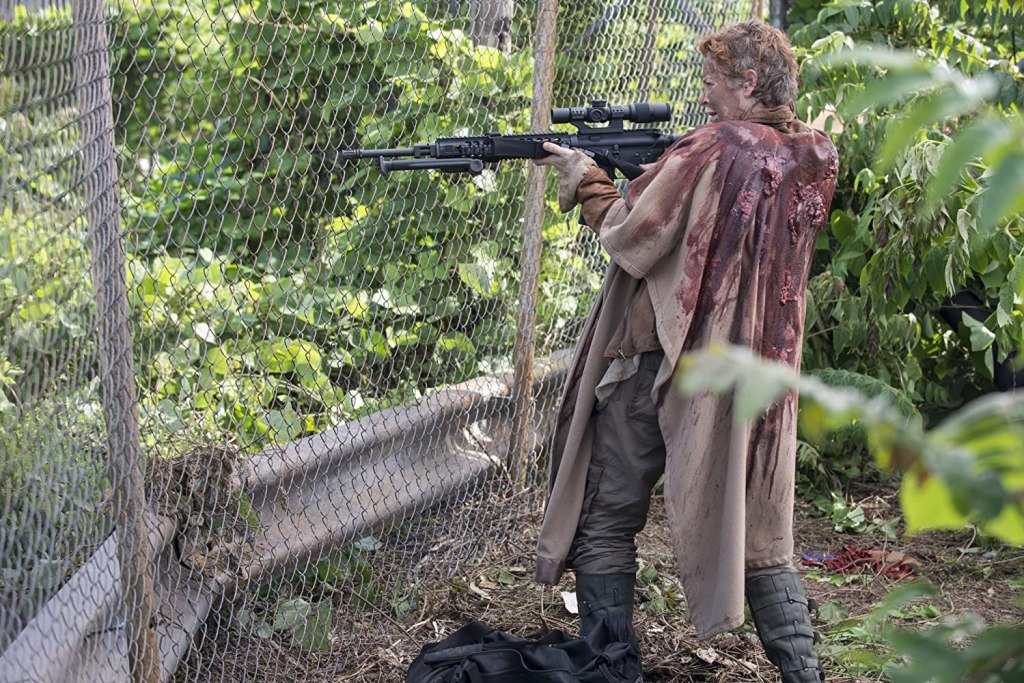 Image still from The Walking Dead episode No Sanctuary. Dressed in a bloodstained cloak, Carol aims a sniper rifle through a steel fence.
