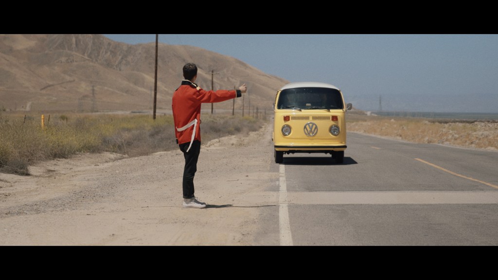 A man in a red coat attempts to hitch a ride from a yellow campervan.