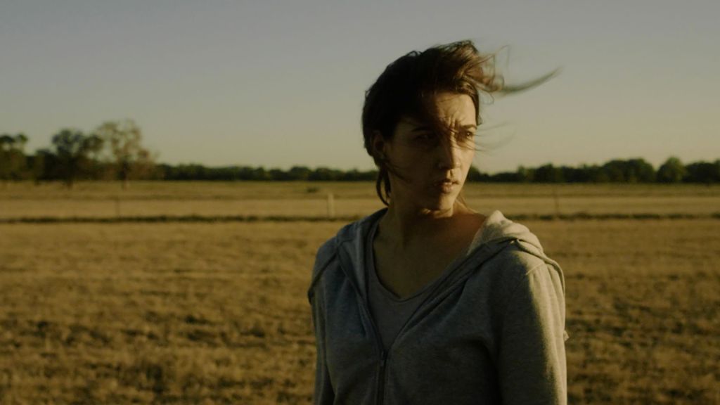 A woman looks distressed and confused in a field. She is wearing a hoodie and t-shirt and her hair blows to the right in the wind.