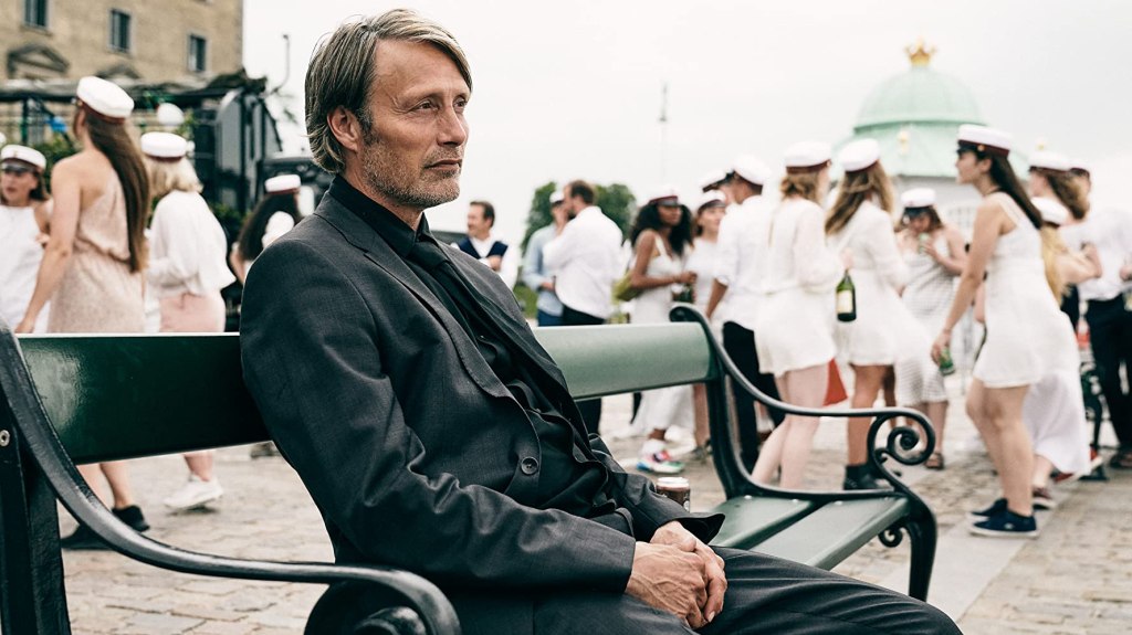 Martin (Mads Mikkelsen), in a dark suit, sits on a bench whilst students in white graduation outfits mingle behind him. From 'Another Round'
