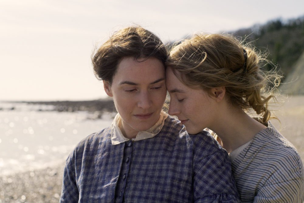 A still from the film Ammonite. Two women walk along the beach wearing period dresses. One is resting her head on the other's shoulder.