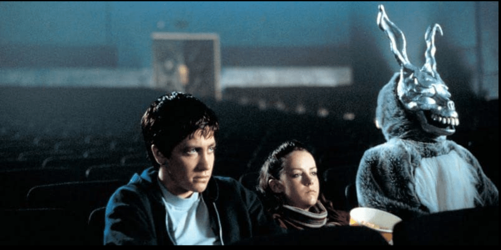 A man, woman, and person in a large rabbit suit with a creepy metal face and ears sit in a movie theater facing the screen