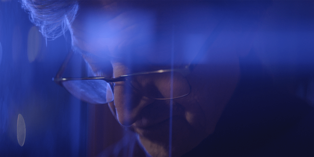 A still from the film Don't Think Twice of an elderly man looking down. He is covered by dark blue lens flares.