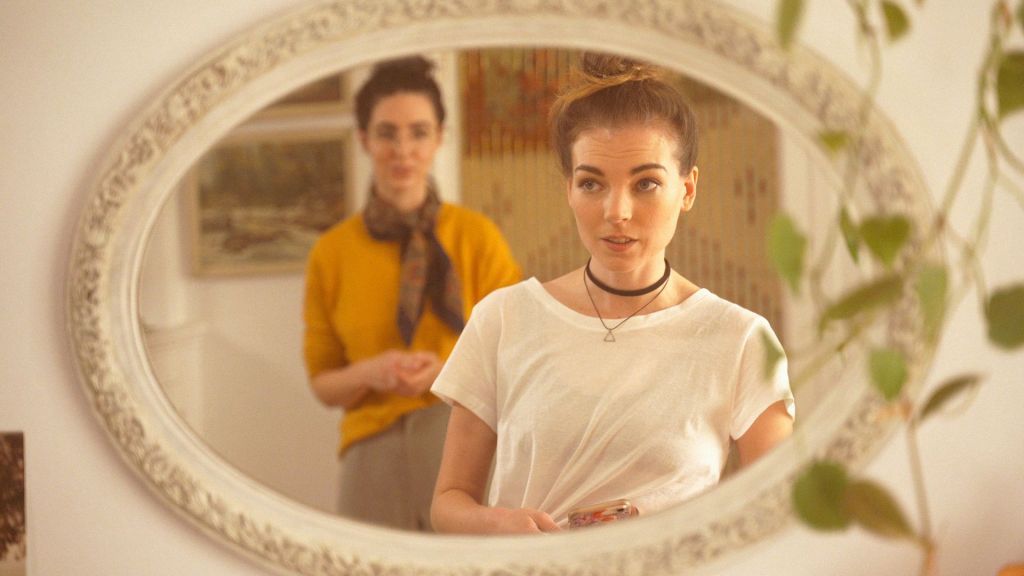 Two women can be seen reflected in an ornate mirror, with one of them being out of focus. The room surrounding them has a "hipster" vibe.