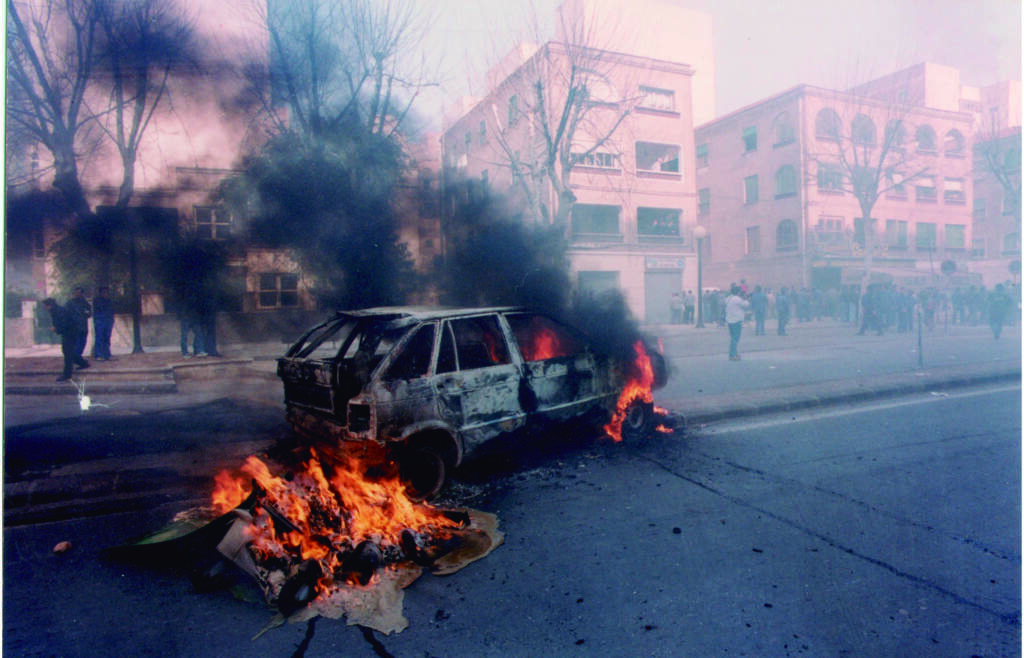 Image from the film 'The Year of Discovery'. A car is on fire in a city street, with a large gathering of people stood away from it.