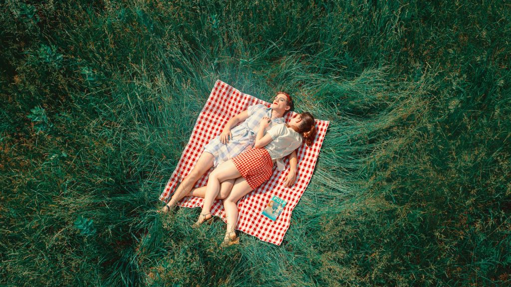 Against green grass, a woman and man lay on a red and white checkered picnic blanket. The man is dressed in drag, with a blue checkered dress, red lipstick, and blue eyeshadow.