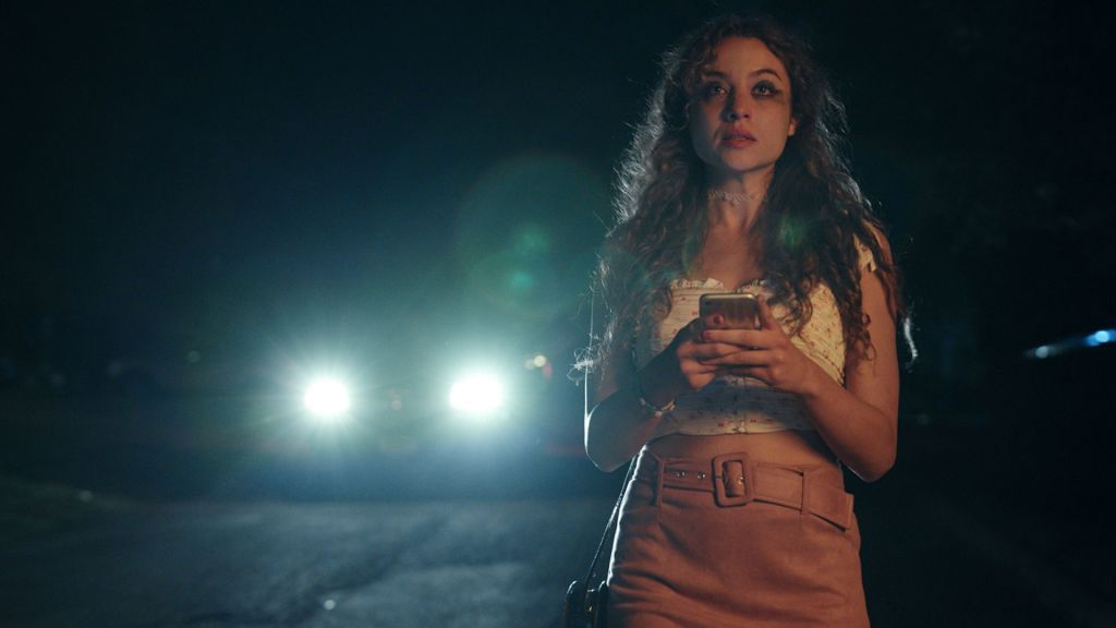 A woman looks past the camera, walking alone at night with car headlights approaching her. She is holding her phone and has smudged mascara on her cheeks.