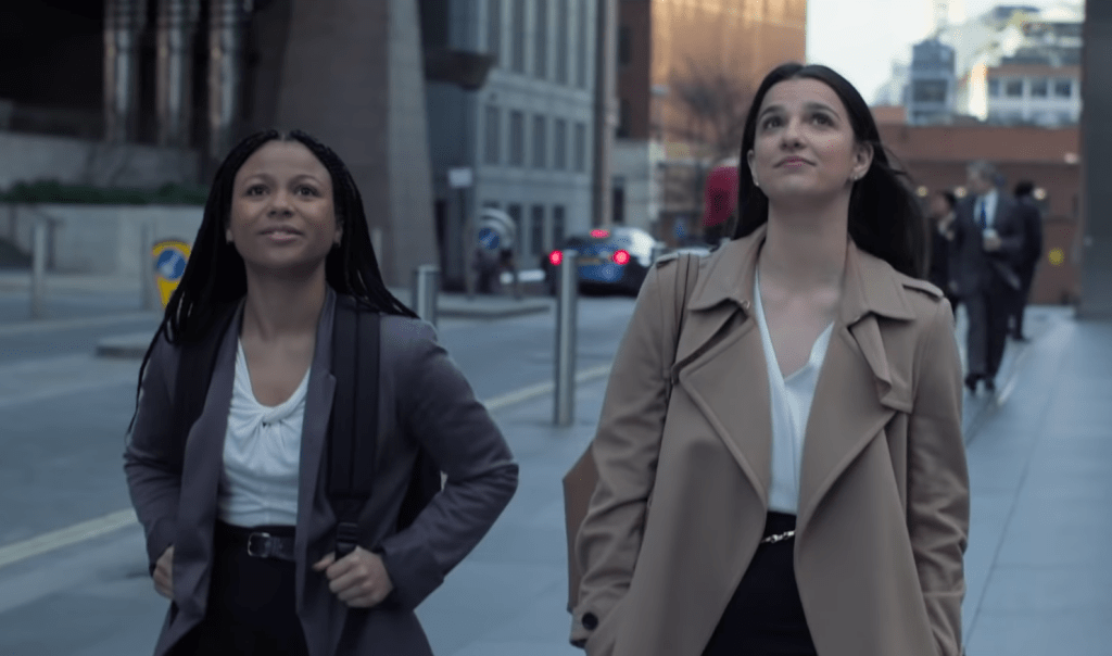 Two women (Myha'la Herrold and Marisa Abela) in business attire walk down a street in a financial distrcit of London, looking up towards something. From 'Industry'