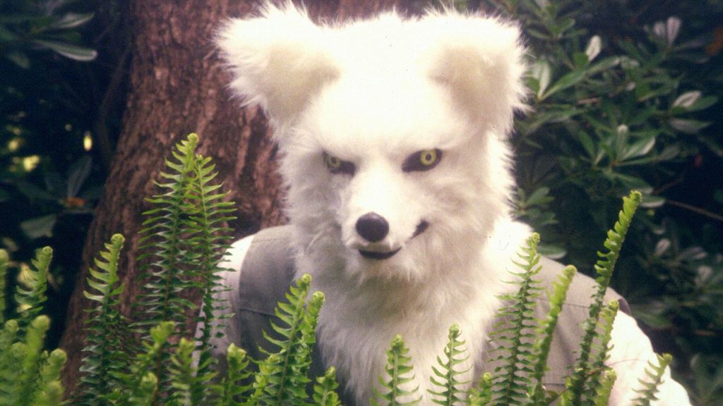 A person in a white fox mask peers from behind a collection of plants. Behind the person is a tree and more greenery.