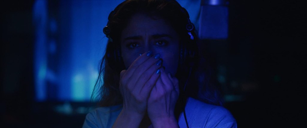 Image still from the horror film The Intruder. A dark, blue-tinted background, a woman (Érica Rivas) dubs sound effects into a microphone.
