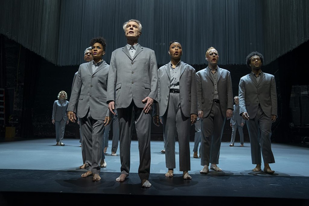 David Byrne and others stand and sing together on stage.