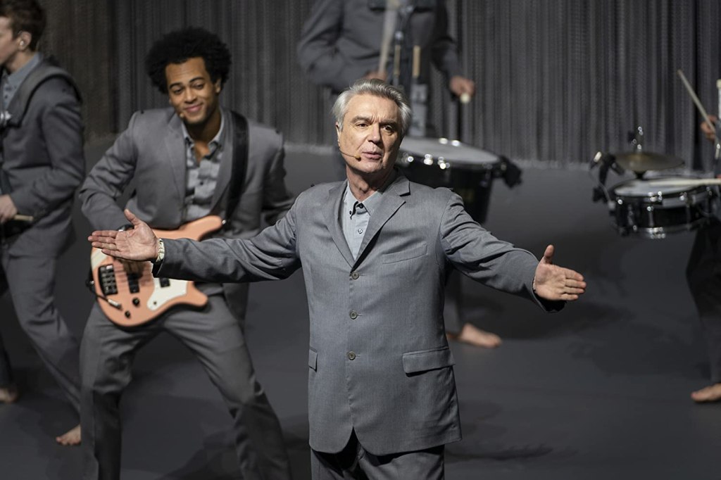 David Byrne sings on stage whilst a musician smiles behind.